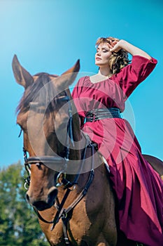 A young woman sits in a red dress is sitting on the back of a brown horse