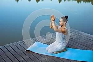 A young woman sits relaxed on a mat by the lake with her folded hands raised above her head. Meditation, yoga in nature