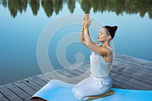 A young woman sits relaxed on a mat by the lake with her folded hands raised above her head. Meditation, yoga in nature