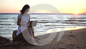 A young woman sits on a stone on the beach by the sea with a laptop in her hands. A girl in a white dress at sunset is