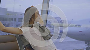 Young woman sits and stretches at airport with airplane on the background