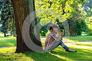 Young woman sits under a tree with a phone in hands in the summe