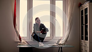 Young woman sits on a window at home with an online tablet. Home interior. 20-25 years