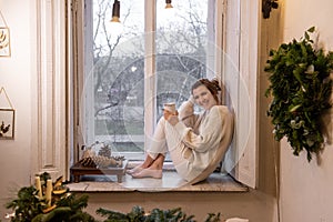 Young woman sits on windowsill near window, holds white cup laughs. Green hand-made Christmas wreath