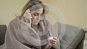 Young woman sits wrapped in a blanket and measures the temperature. She has symptoms of flu, cold or coronavirus.