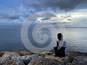 Young woman sitting alone on the beach watching the sea view. Blue sky over the ocean horizon. Tranquil nature scenery. Self love