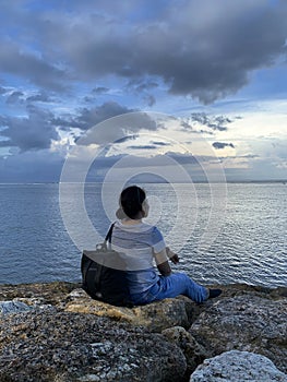 Young woman sitting alone on the beach watching the sea view. Blue sky over the ocean horizon. Tranquil nature scenery. Self love