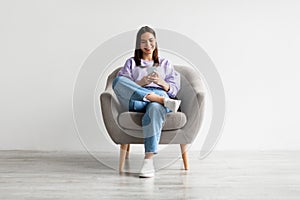 Young woman sitting in armchair, looking at mobile phone screen, using new cool application against white wall