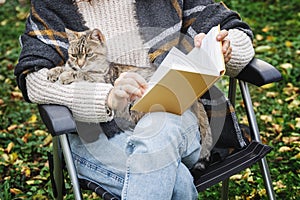 Young woman sitting in the autumn garden with a pet gray cat reading a book.