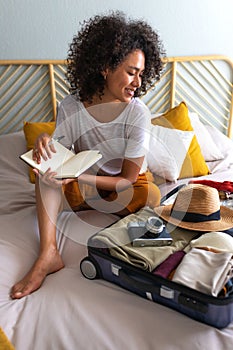 Young woman sitting on bed checking packing list before summer holidays. Preparing suitcase for vacations. Vertical