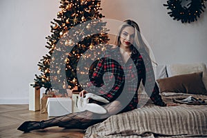 Young woman sitting on bed and holding bunny mask against glowing Christmas tree