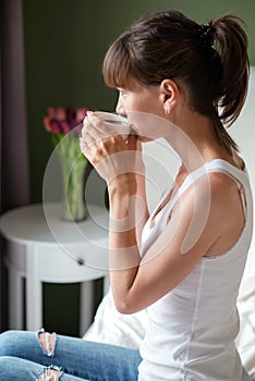 Young woman sitting on the bed, holding a white cup and looking out the window