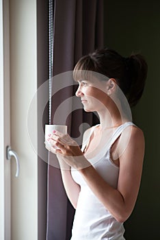 Young woman sitting on the bed, holding a white cup and looking out the window