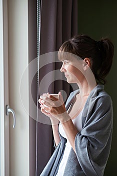 Young woman sitting on the bed, holding a white cup and looking out the window