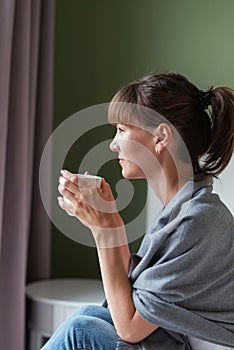 Young woman sitting on the bed, holding a white cup and looking out the window