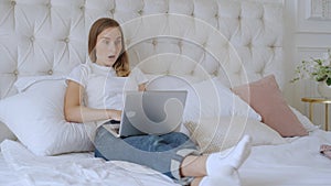 Young woman sitting on the bed with laptop cheering carefree and excited. Victory concept.