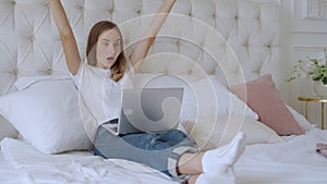 Young woman sitting on the bed with laptop cheering carefree and excited. Victory concept.