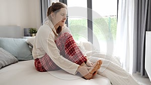 Young woman sitting in bed and massaging her aching feet because of muscle cramp