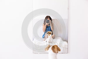 young woman sitting on bed and taking a picture with a reflex camera to her cute small dog on the mirror. Daytime. LIfestyle with