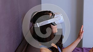 Young woman sitting on the bed and using VR headset, playing game and pointing in the air. Lady in bedroom at home