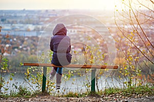 Young woman sitting on bench in autumn city park and looking at cityscape, back view