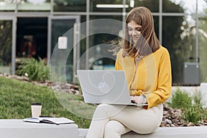 Young woman is sitting on a bench of city park and using laptop computer. Focused european business woman student