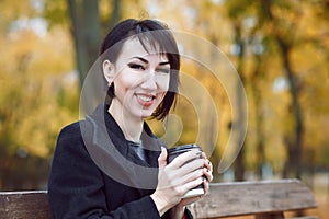 Young woman sitting on a bench and drinking coffee, autumn season, city park, yellow leaves