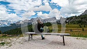 Young woman sitting at bench, drinking glass of red wine, looking at Cima Ambrizzola in front of Rifugio Cinque Torri. Dolomites, photo