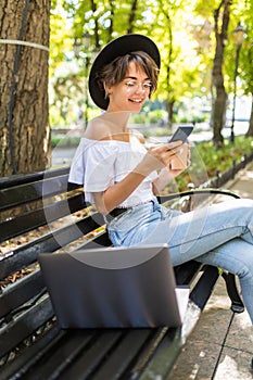 Young woman sitting on bench in green park on summer day and reading text message on cell phone while using silver laptop