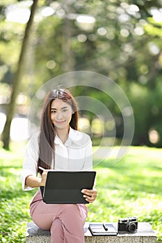 Young woman sitting on bench in the park with digital tablet and smiling to camera.