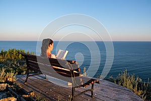 Young woman sitting on a bench and reading book outdoors with summer sea background