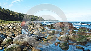 Young woman sitting on a big rock on the Baltic sea coast. Redhead woman relaxing on the seashore