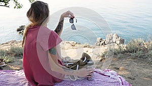 Young woman sitting on a blanket on the beach by the sea, looking in front of her and holding a thermos cup, travel mug