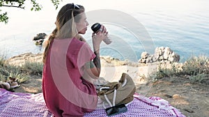 Young woman sitting on a blanket on the beach by the sea, looking in front of her and holding a thermos cup, travel mug