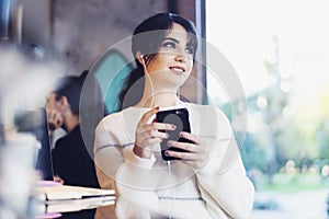 Young woman sitting cafe at table, thoughtfully looking out window, holding smartphone.Portrait girl with cell phone in her hands