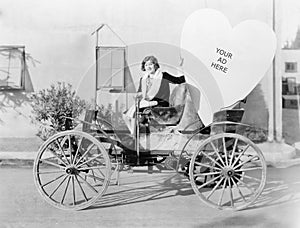 Young woman sitting on a car holding a big heart shaped sign