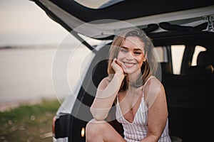 Young woman sitting in a car trunk near lake, enjoying summer time.