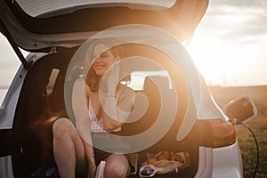 Young woman sitting in a car trunk, waiting for charging.