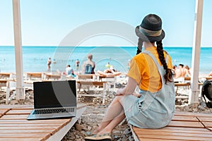 A young woman is sitting on a chaise longue with a laptop next to her. Beach and sea in the background. Rear view. The concept of