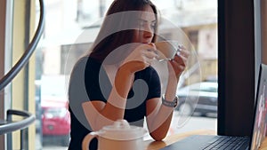 Young woman sitting in the coffee shop by the window having a coffee break, she is using her laptop, working and surfing