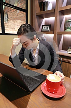 Young woman sitting in coffee shop at wooden table, drinking coffee and thinking problem of anxiety.On table is laptop