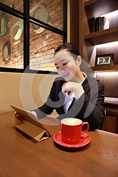 Young woman sitting in coffee shop at wooden table, drinking coffee and using pad.On table is laptop