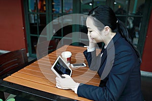 Young woman sitting in coffee shop at wooden table, drinking coffee and using pad. On table is laptop