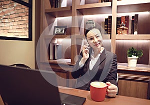 Young woman sitting in coffee shop at wooden table, drinking coffee and using smartphone.On table is laptop