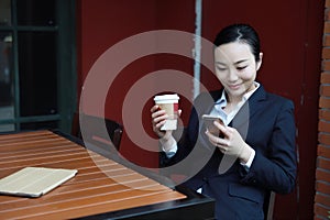 Young woman sitting in coffee shop at wooden table, drinking coffee and using smartphone.On table is laptop