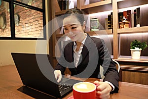 Young woman sitting in coffee shop at wooden table, drinking coffee and using smartphone.On table is laptop