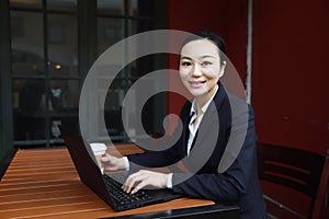 Young woman sitting in coffee shop at wooden table, drinking coffee and using smartphone.On table is laptop