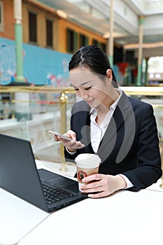 Young woman sitting in coffee shop at wooden table, drinking coffee and using smartphone.On table is laptop