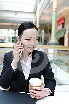 Young woman sitting in coffee shop at wooden table, drinking coffee and using smartphone. On table is laptop