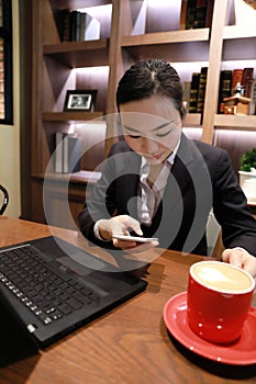 Young woman sitting in coffee shop at wooden table, drinking coffee and using smartphone. On table is laptop